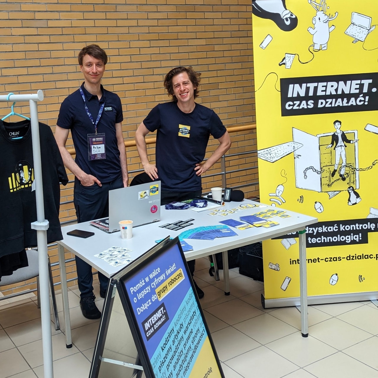 Zdjęcie stoiska Fundacji „Internet. Czas działać!” na Poznańskiej Imprezie Wolnego Oprogramowania. Widać dwie osoby stojące za stolikiem, na którym stoi laptop oraz leżą ulotki i naklejki. Po lewej stronie widać stojak z koszulkami, po prawej żółto-czarny rollup z logo Fundacji. Przed stolikiem widoczny potykacz z plakatem informującym o grupie roboczej Fundacji.