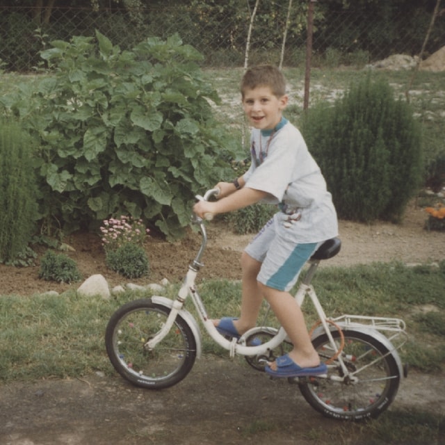 Zdjęcie kilkuletniego Mateusza Jarczyńskiego na białym dziecięcym rowerze. Ma na sobie koszulkę oraz krótkie spodenki. W tle widać rabatę kwiatową w ogrodzie.