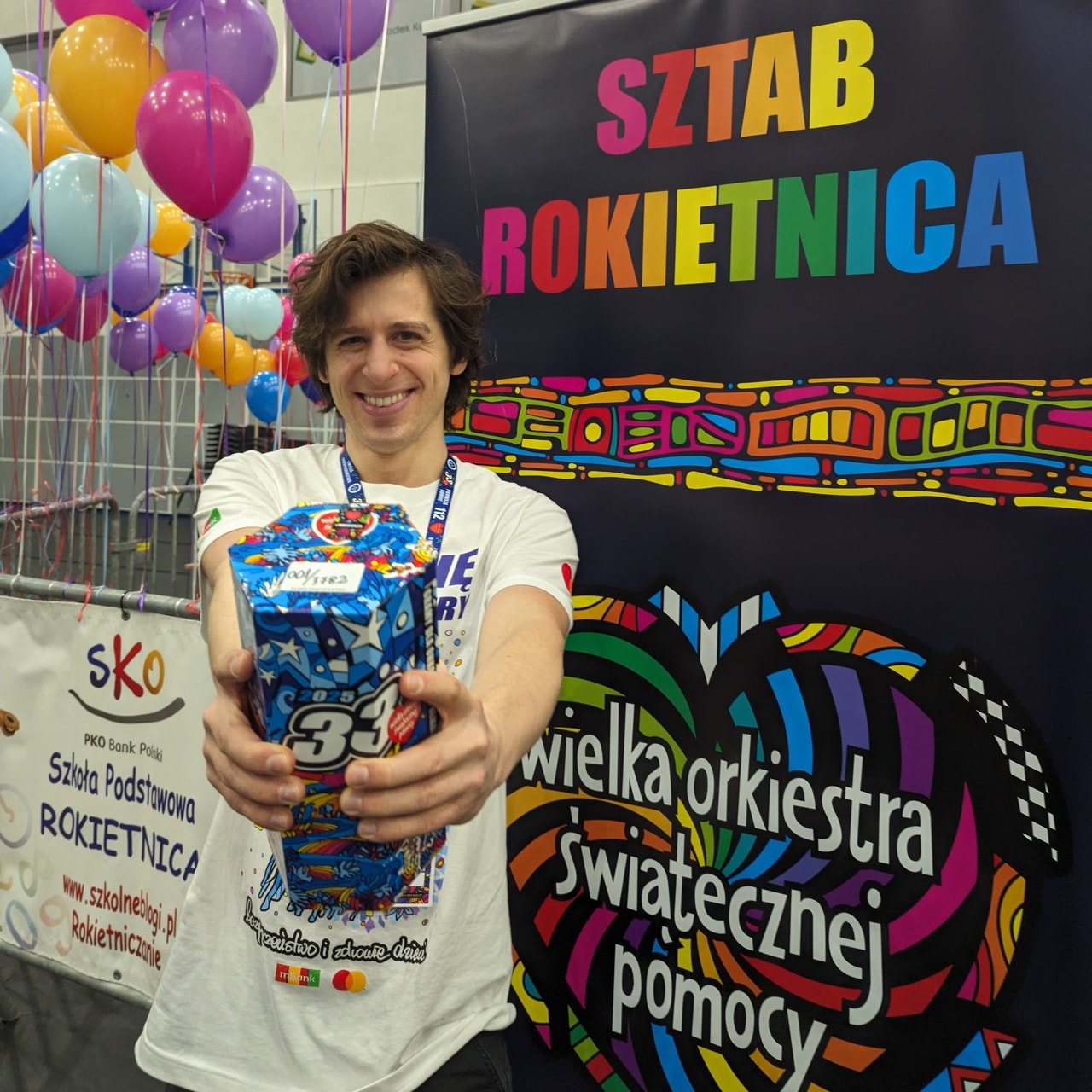 Zdjęcie Mateusza Jarczyńskiego z jako wolontariusza Wielkiej Orkiestry Świątecznej Pomocy. W wyciągniętych przed siebie rękach, na pierwszym planie, trzyma papierową puszkę na datki. Ma na sobie białą koszulkę z grafikami Finału WOŚP. Po prawej widoczny kolorowy rollup z logo WOŚP oraz tęczowym napisam „Sztab Rokietnica”.