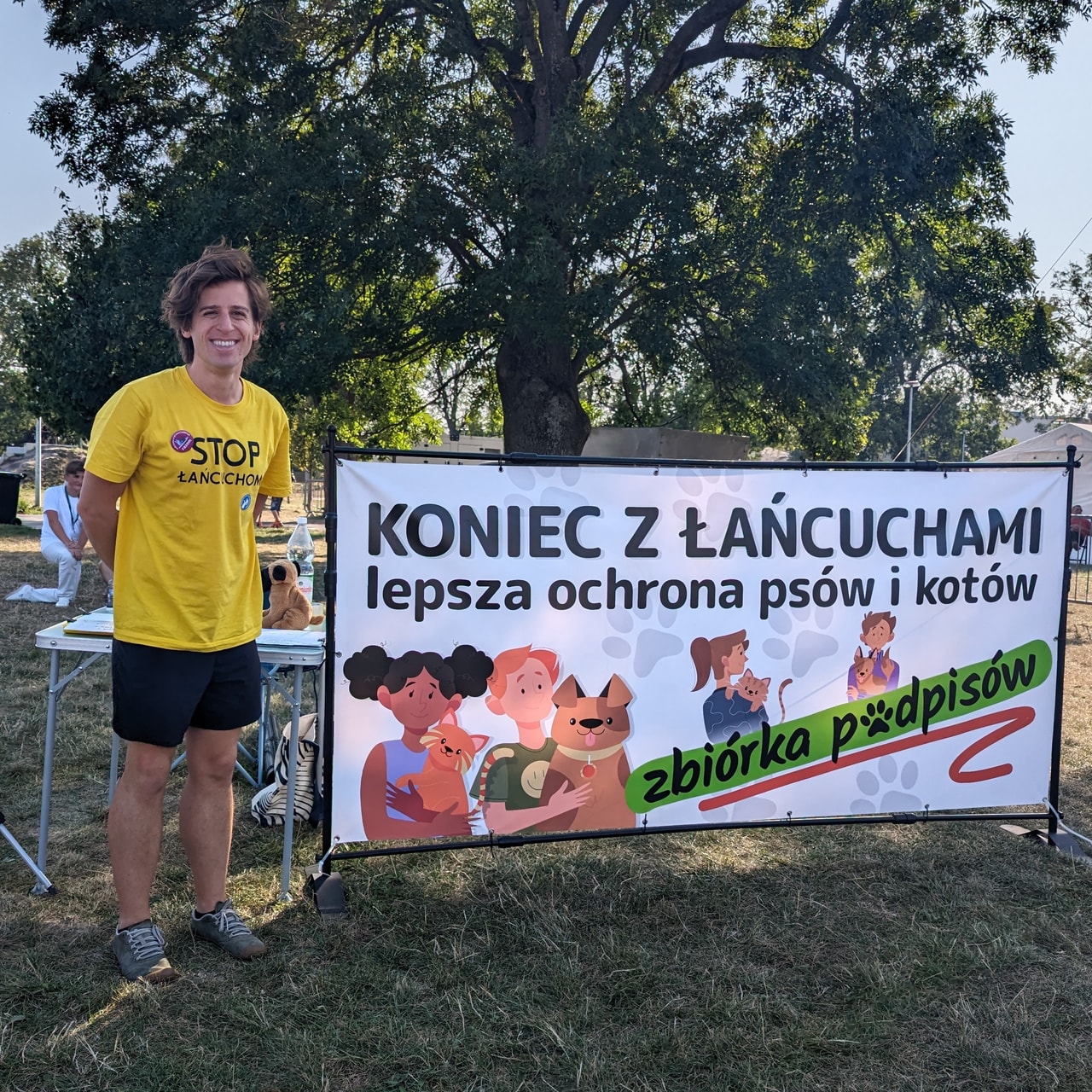 Zdjęcie Mateusza Jarczyńskiego w żółtej koszulce z napisem „Stop łańcuchom” przy stoliku do zbiórki podpisów oraz banerze z napisem „Koniec z łańcuchami. Lepsza ochrona psów i kotów. Zbiórka podpisów” oraz grafikami przedstawiającymi ludzi trzymających w rękach zwierzęta domowe.